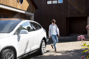 A man carrying a charging cable walks away from his white electric vehicle parked in front of a wooden house