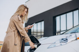 A woman in a beige trench coat uses her phone while plugging in her electric vehicle at a charging station in front of her condo.